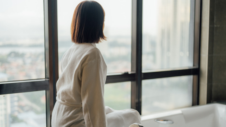 Woman sitting on bathtub looking at a large uncovered window