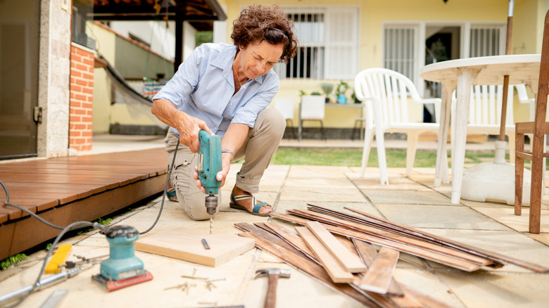 A person drilling wood in a backyard beside an outdoor wood deck and nearby buildings