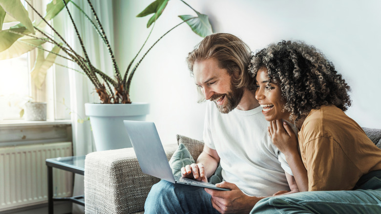 A smiling couple sits on a sofa while looking at homes online