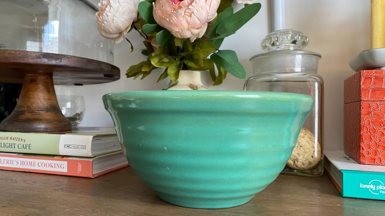 A green Bauer pottery bowl on a counter with cookbooks, a flower vase, cake stand, and cookie jar