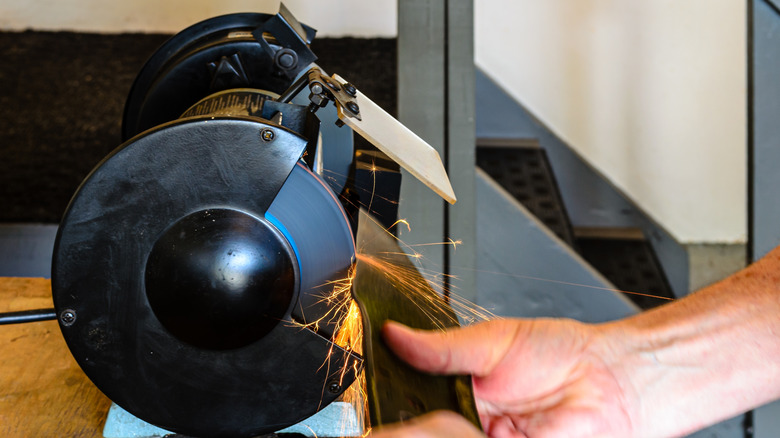 Hands sharpening a lawn mower blade against a bench grinder