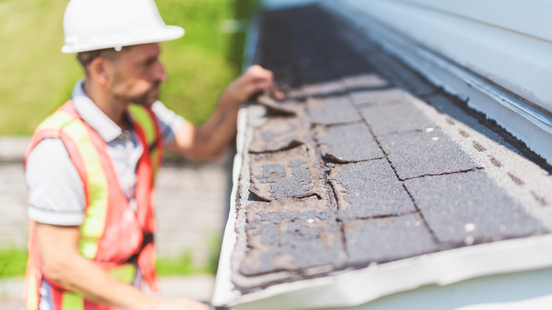 A man wearing a white hard hat inspects damaged shingles on a residential roof
