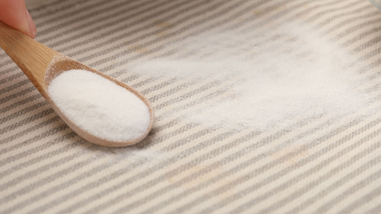 A person using baking soda in a wooden spoon on couch fabric to reduce odors.