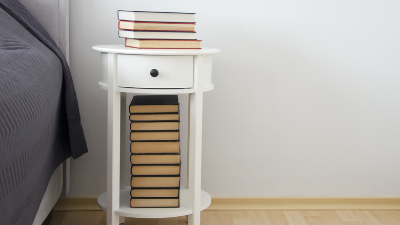 stack of books on a white nightstand beside a bed in a neutral colored room