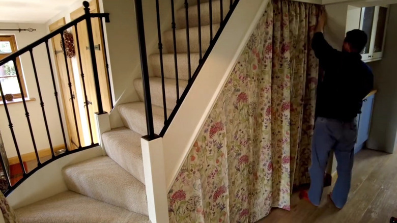 A man hanging a long curtain under his stairs.