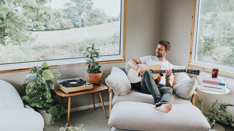 Man in comfy chair playing guitar, sitting in corner with big windows