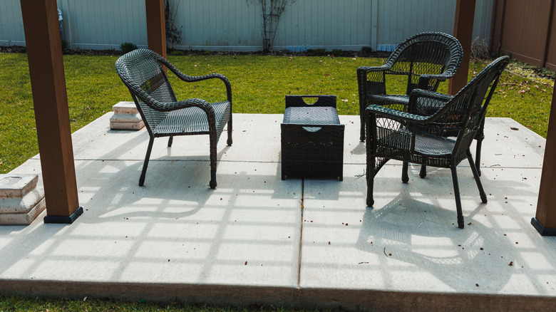 A concrete patio with black wicker chairs and a table in front of a green yard