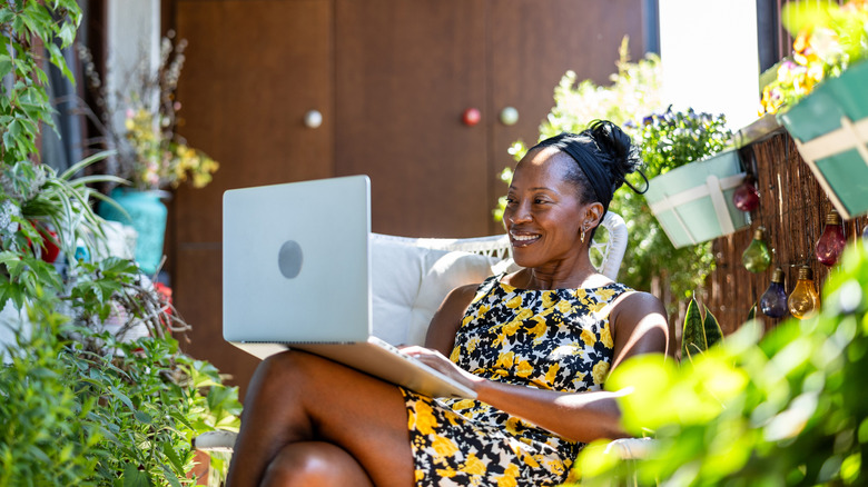A woman smiles while looking at her laptop and sitting on her patio in a colorful dress