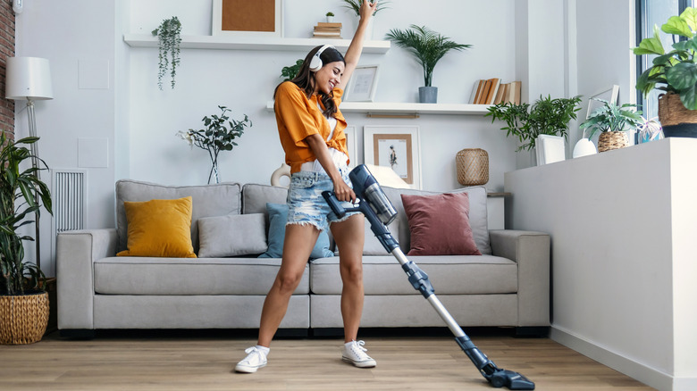 A woman in headphones dances while using a cordless stick vacuum on her bare floors.