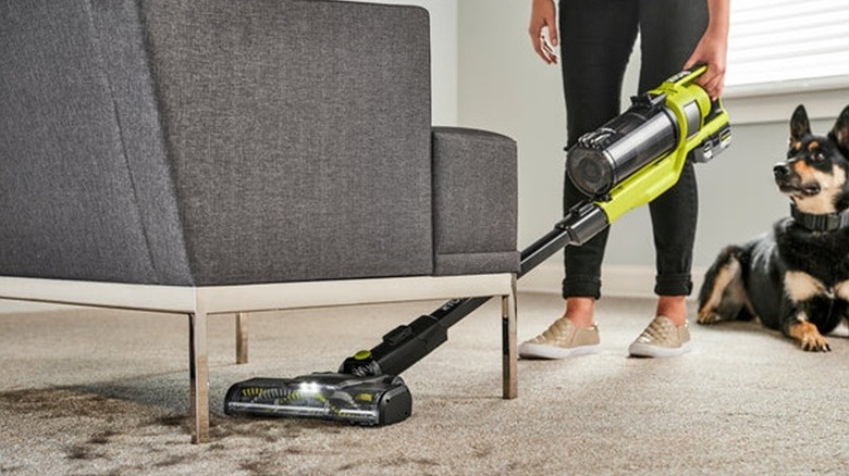 A woman uses the Ryobi One+ HP stick vacuum on pet hair under a gray chair, with a dog looking on.