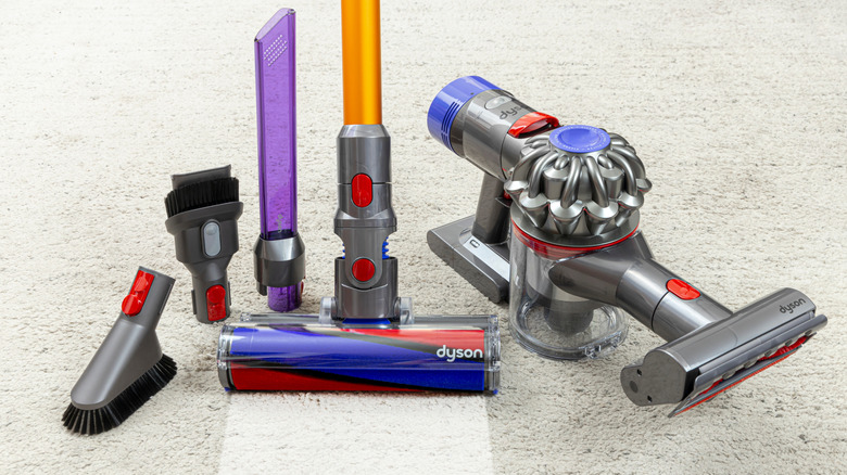 A handheld and cordless stick Dyson vacuum sits on carpeting with all of its attachments