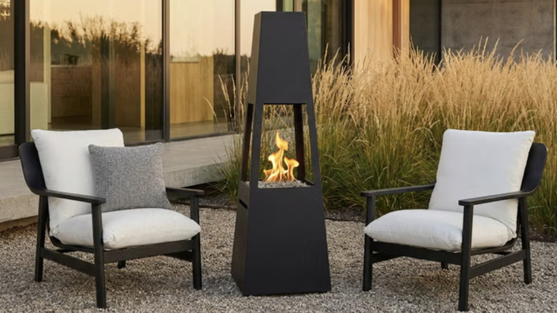 Real flame patio set with modern chairs, a chiminea fire pit in a modern backyard with tall grass in the background.