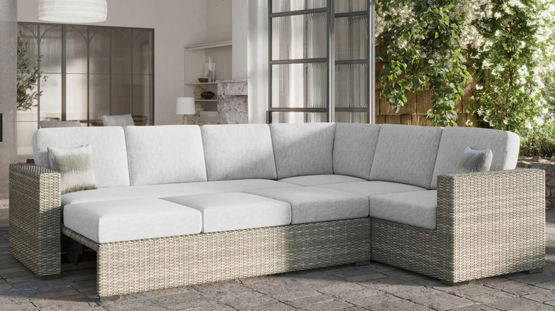 A light wicker and gray cushioned outdoor sectional sits on a backyard patio