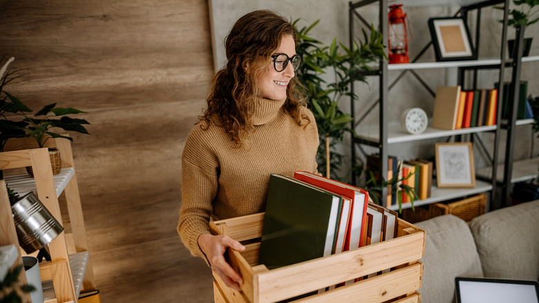 A woman with glasses in a living room holding a wooden storage bin with books