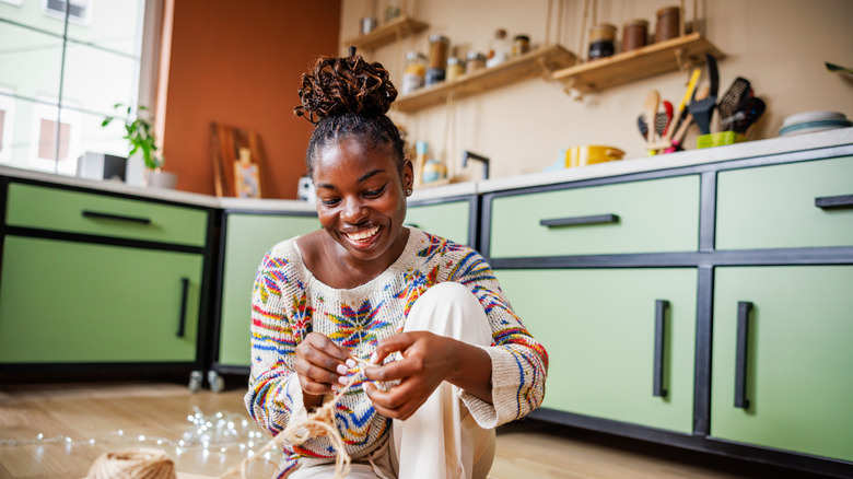A woman sits on the floor of a green kitchen, smiling as she holds twine for a craft