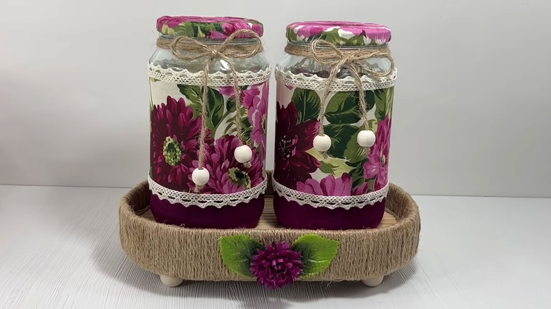 Two old jars covered in matching purple fabric and ribbon sit inside a jute and wood tray