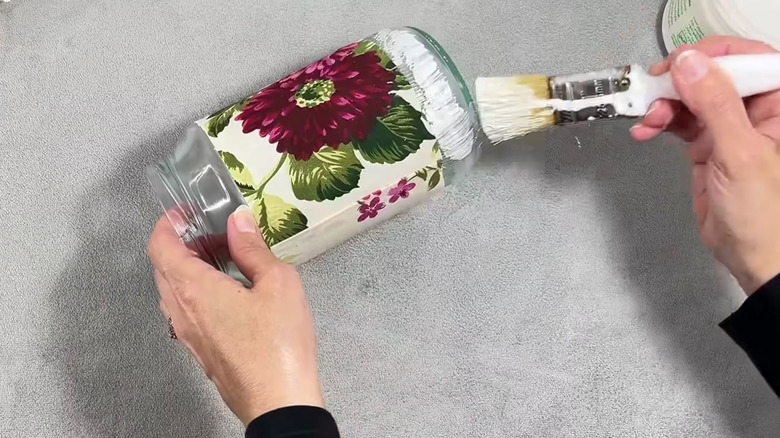 A person applies ModPodge to an old jar with a paintbrush