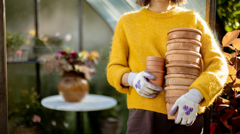 A person in a bright yellow sweater and floral gardening gloves holds a stack of terracotta pots