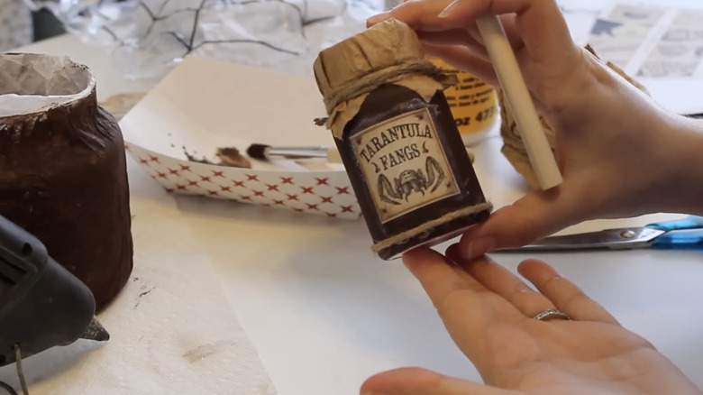 A diy potion bottle in brown with rustic touches; a woman is holding it over a table where she is crafting