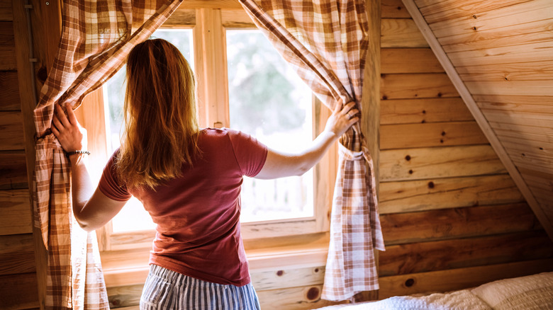 A woman in a red t-shirt opening gingham blinds in a wooden cabin