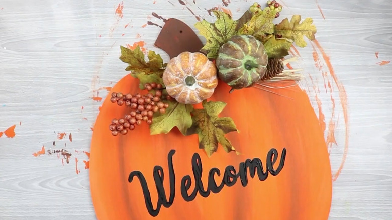 A pumpkin-shaped door sign with the word Welcome painted on, decorated with faux pumpkins and leaves