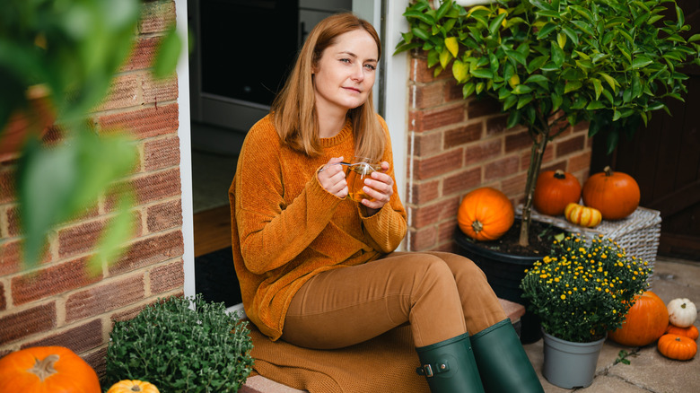 A woman sits by her front door on a fall day, surrounded by pumpkins