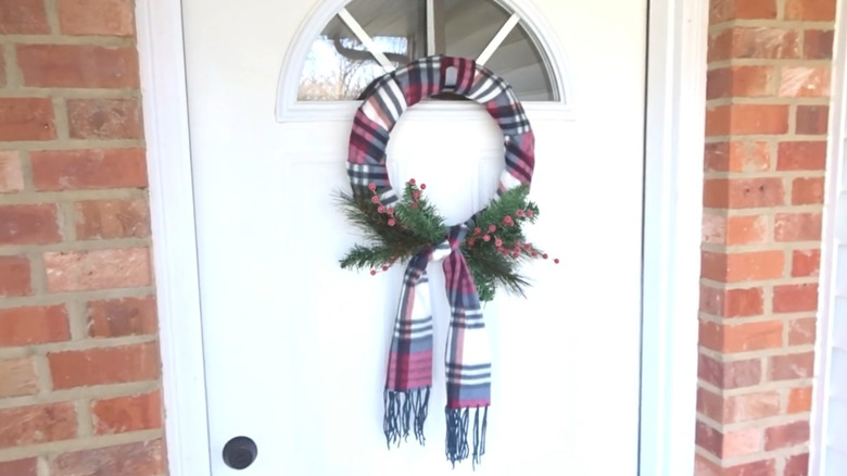 A white front door with a wreath made of a scarf, pine branches, and red berries