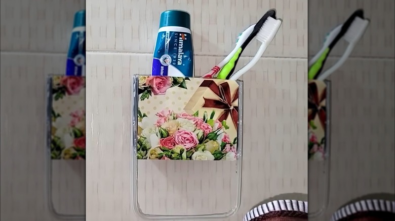 Takeout container repurposed as floral toothbrush and toothpaste holder on bathroom tile wall