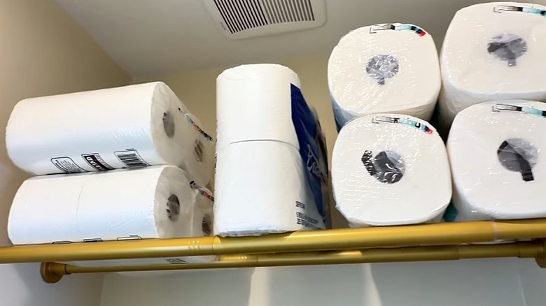Two tension curtain rods holding up rolls of toilet paper and paper towels in a bathroom