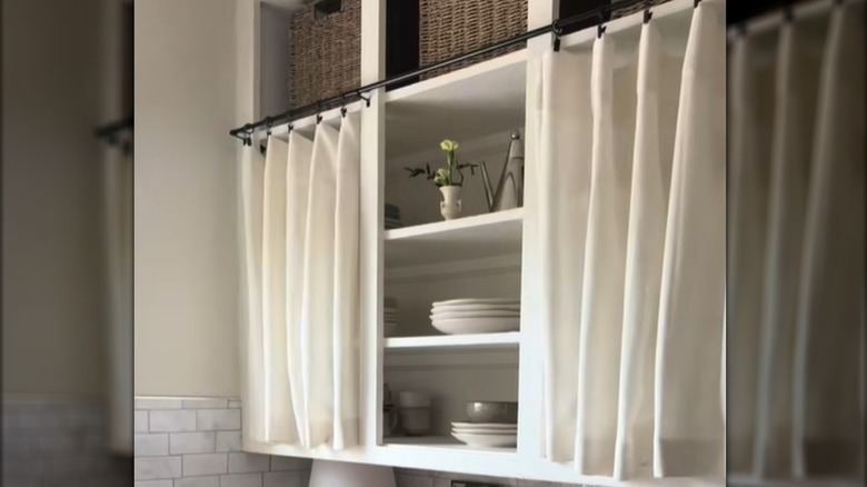 White curtains hung over open-shelf kitchen cabinets with dishes and baskets