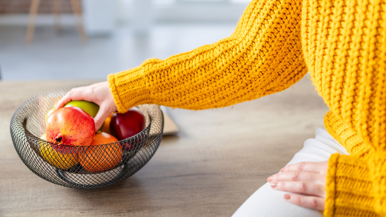 Woman taking apple from full fruit bowl on kitchen table