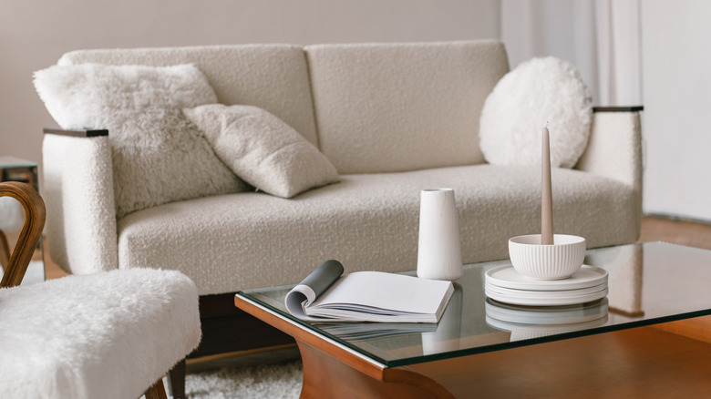 A neutral living room with a white boucle sofa, white imitation-fur chair, white pillows, and white decor on a wood and glass coffee table