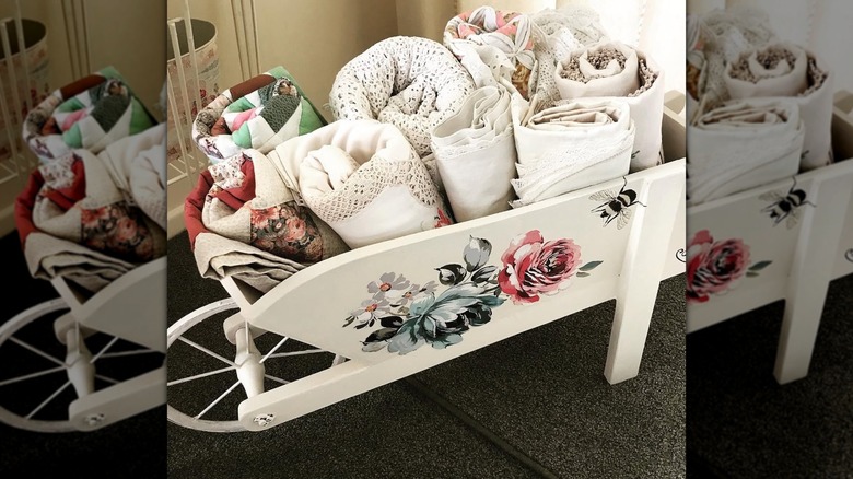 A wooden wheelbarrow painted white adorned with decals and used for blanket storage