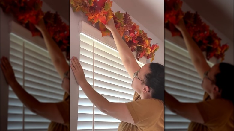 Woman hanging pool noodle garland with fall leaves above house windows