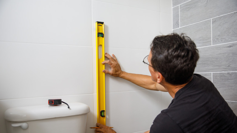 Man using level to mark wall beside toilet tank in white and gray bathroom