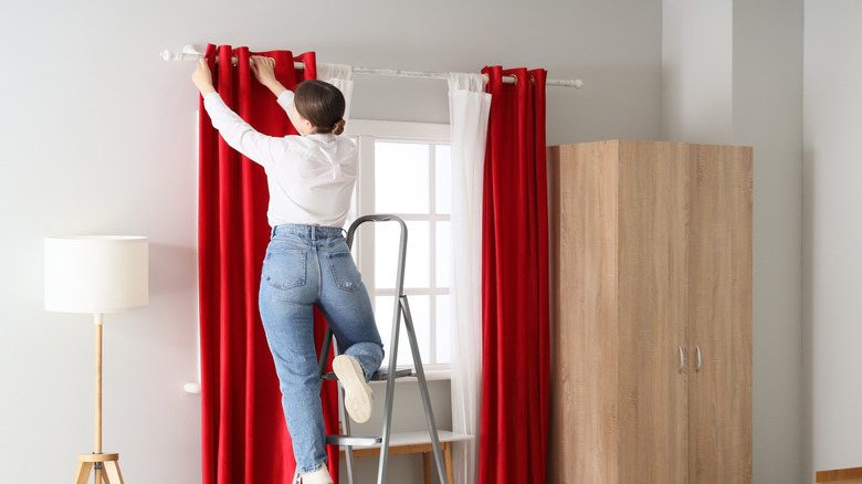 A woman hangs red curtains from a rod.