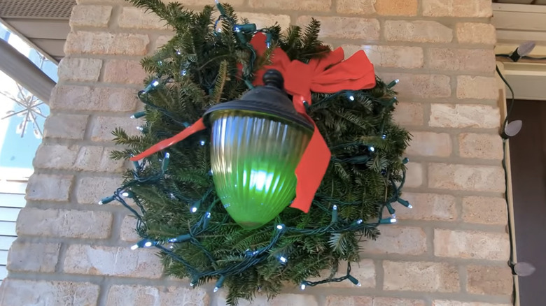 A holiday wreath around a porch light with a green bulb inside