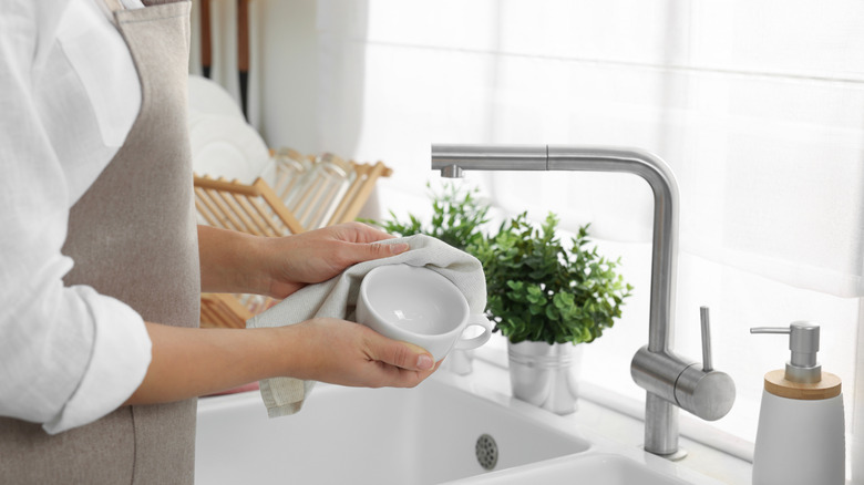 A woman wiping a white coffee mug with a white dish towel over a kitchen sink