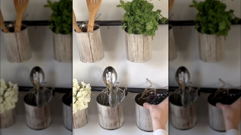 A person tucking kitchen herbs, decorative flowers, and kitchen utensils into a DIY soup can rack