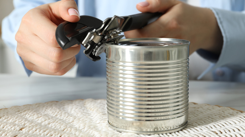 A person opening tin can with can opener over woven placemat on white table