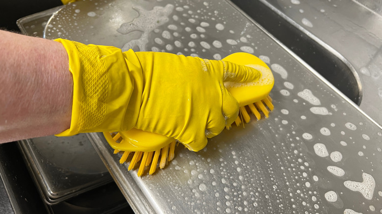 a gloved hand scrubs a baking sheet with a brush