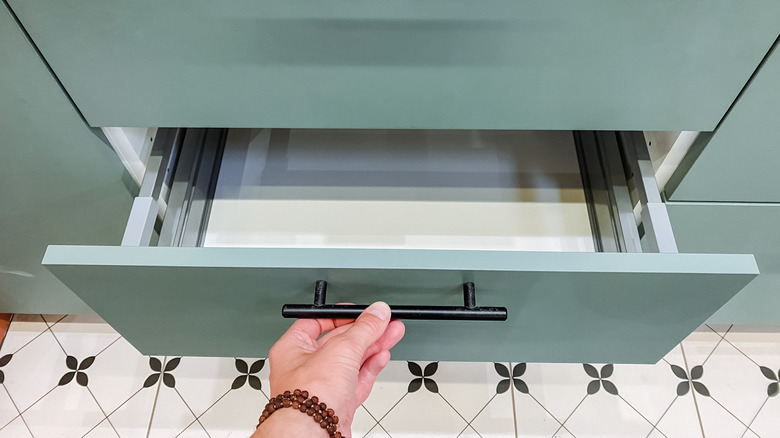 A woman with a bracelet opening an empty green kitchen cabinet lined with a white drawer liner