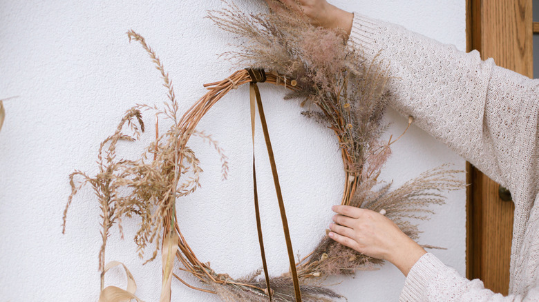 A person taking down a Thanksgiving wreath with grass foliage from a front porch wall.