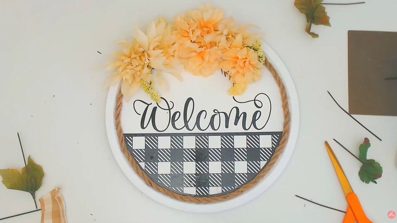 Dollar Tree welcome sign with braided rope, orange flowers, and a plaid pattern on a white worktable
