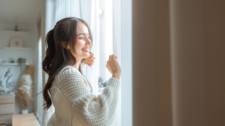 A woman in a knitted cardigan smiles by a window with basic curtains