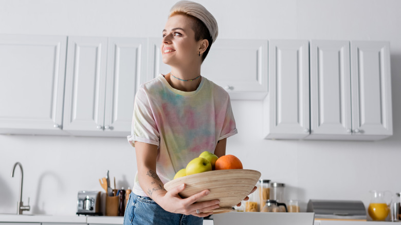 A woman holds a bowl of fruit in a white kitchen