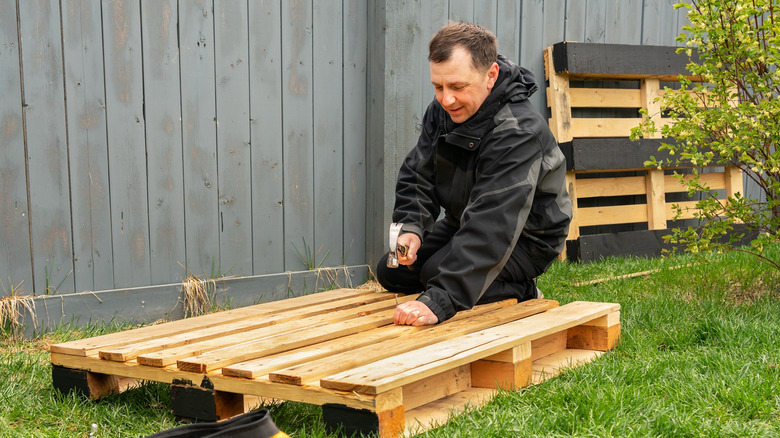 A man in kneels in the grass and uses a hammer and nail to work on a DIY project with a wood pallet