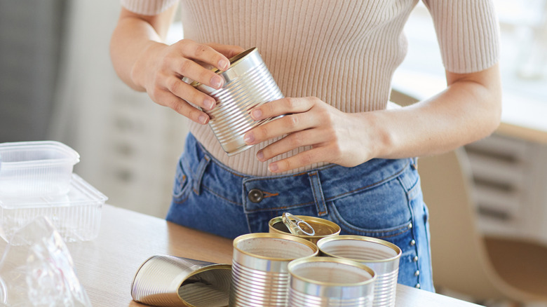 Woman holding tin can over multiple cans on kitchen counter