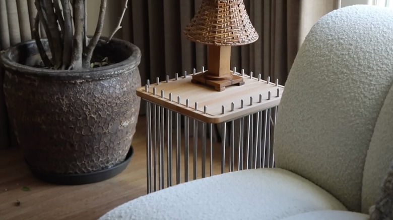 A lamp sits on a modern silver bar and wood side table between a boucle couch and potted plant
