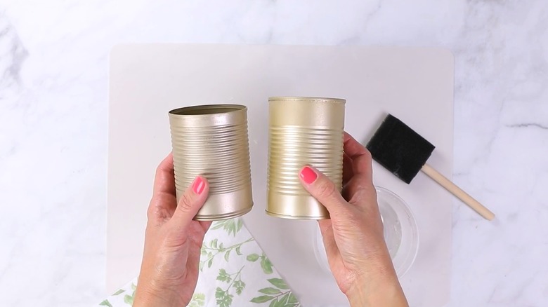 Woman holding up two painted tin cans for DIY project over stone countertop with paper napkins